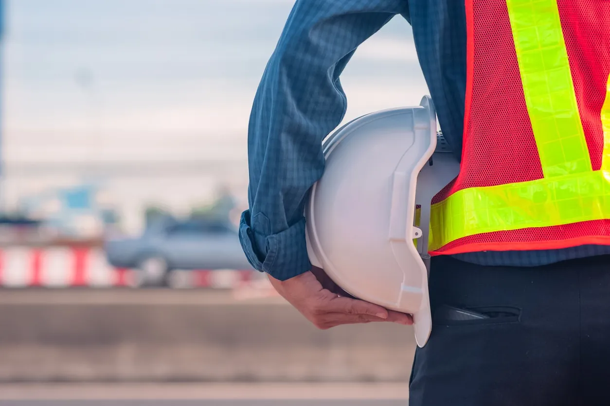 Field worker in a high-visibility vest holding a white hard hat beside a roadway